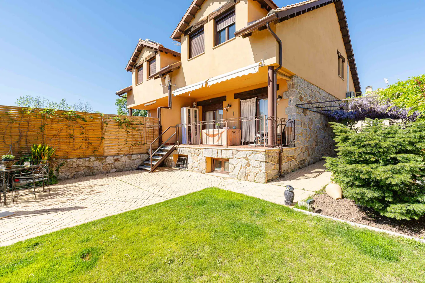Exterior view of a two-story tan house with a stone foundation, a patio, and a green lawn.