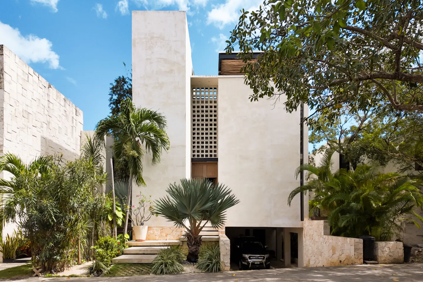 Modern white stucco house with a car parked underneath, surrounded by lush tropical greenery and a blue sky.