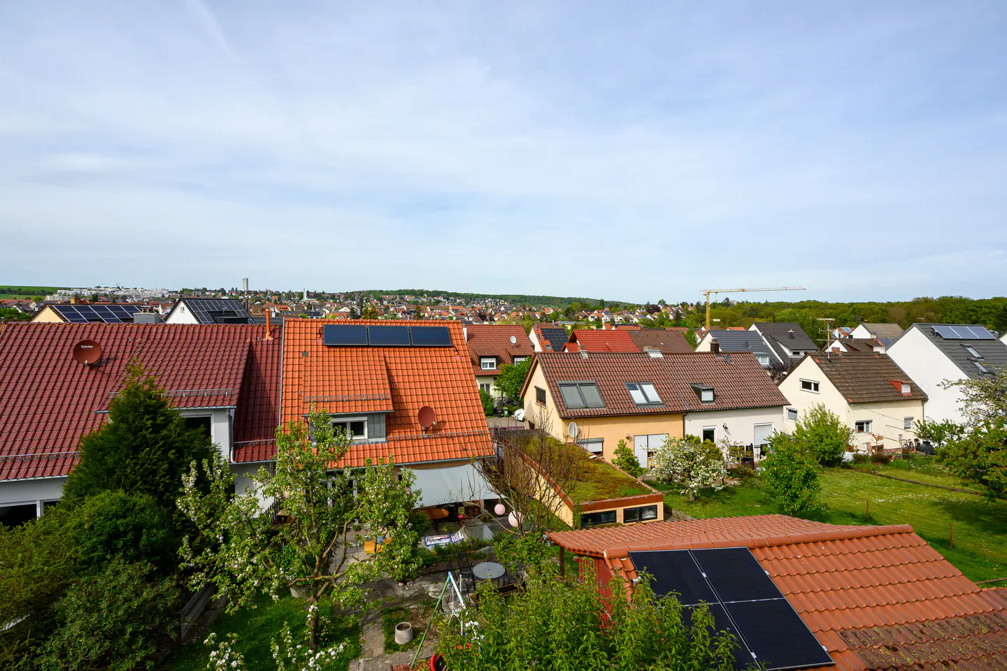 View of a residential neighborhood with houses with red roofs, some with solar panels, and green trees under a blue sky.