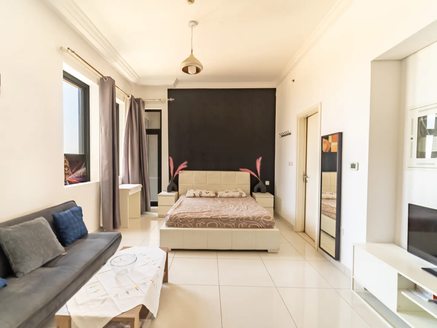 Modern studio apartment with a grey sofa, coffee table, and a queen bed against a black accent wall on white tile floors.