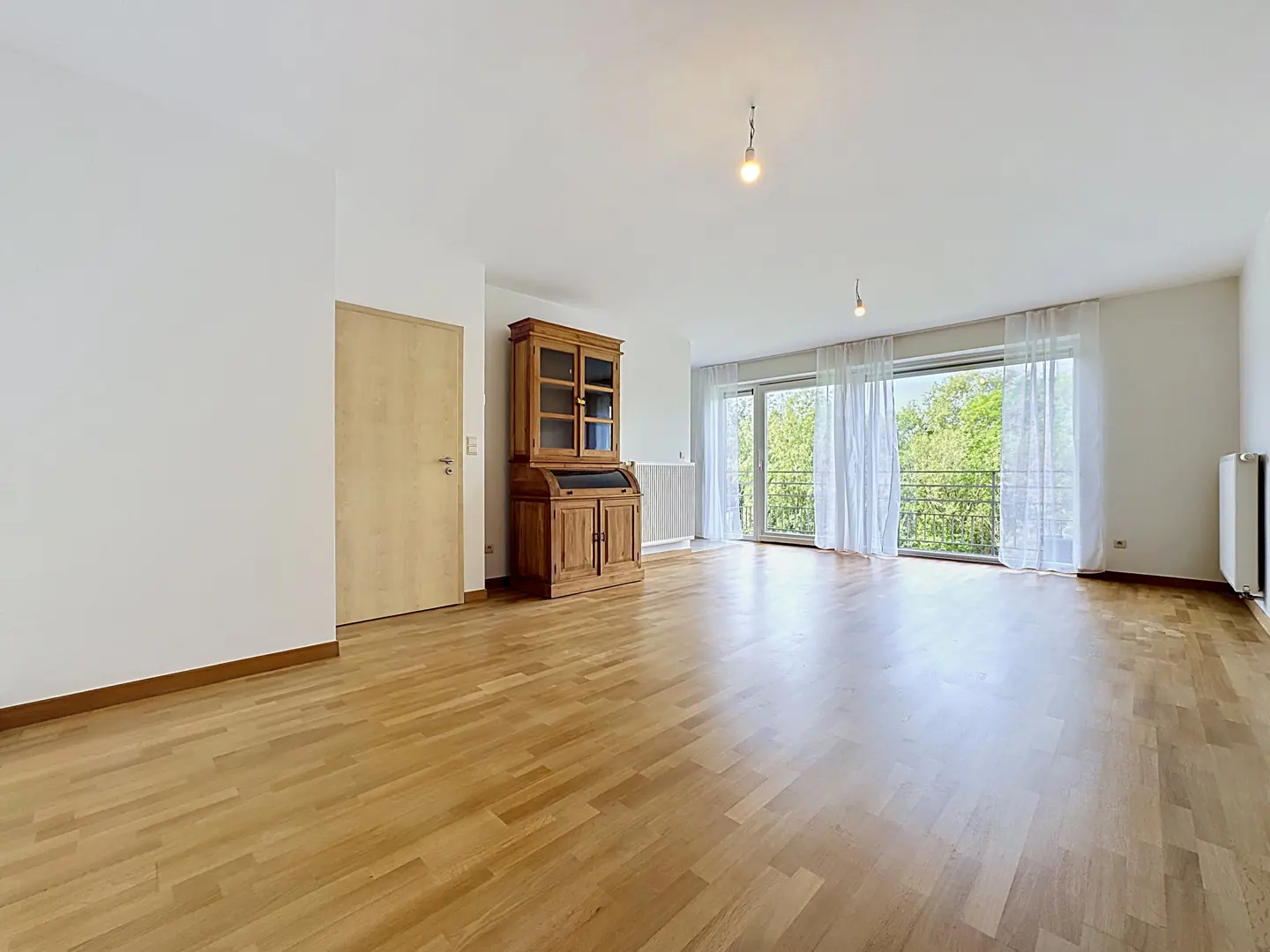 Bright, empty room with wood floors, white walls, and a large window with sheer curtains. A wooden cabinet stands near a door.