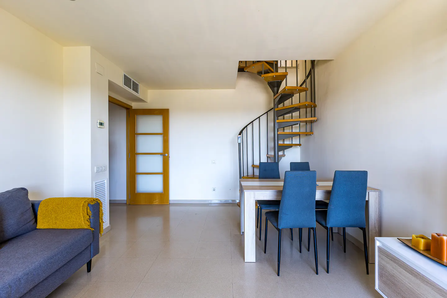 Modern living room with a grey sofa, yellow throw, dining table with blue chairs, and a metal spiral staircase.