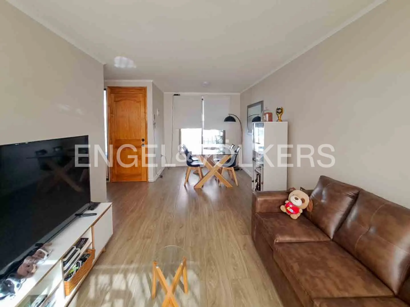 Living room with wood floors, brown leather sofa with a teddy bear, TV, dining table with chairs, and a white shelving unit.