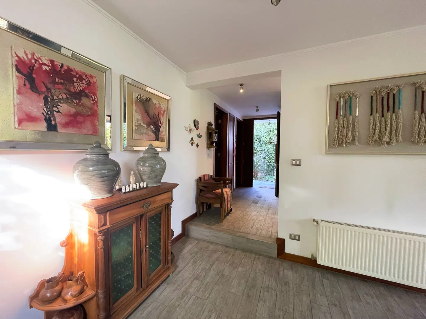 A bright hallway with a wooden cabinet, two large grey vases, abstract art, and a doorway leading to a garden.