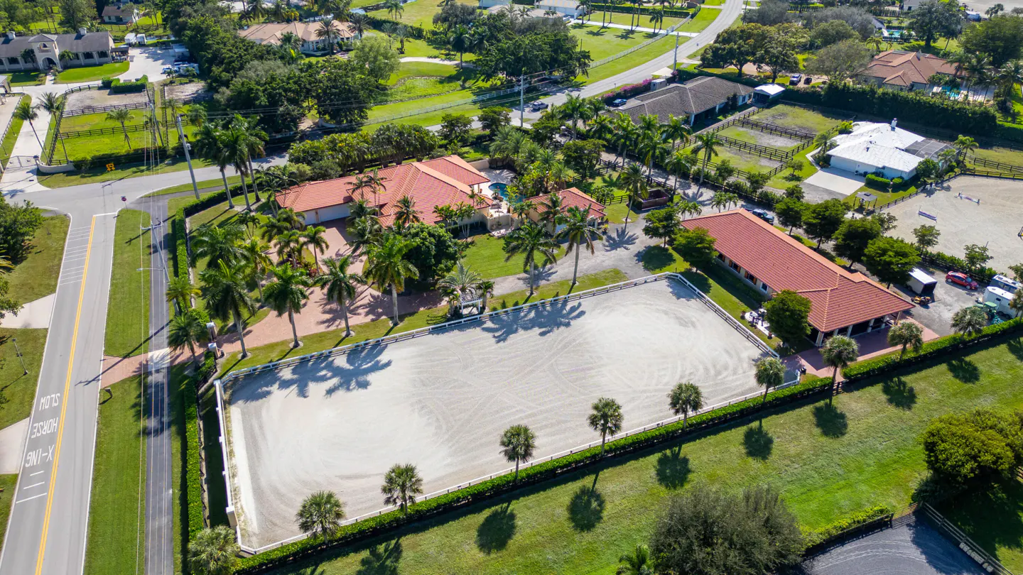 Aerial view of an equestrian estate with a sand arena, red-roofed buildings, palm trees, and lush green landscaping.