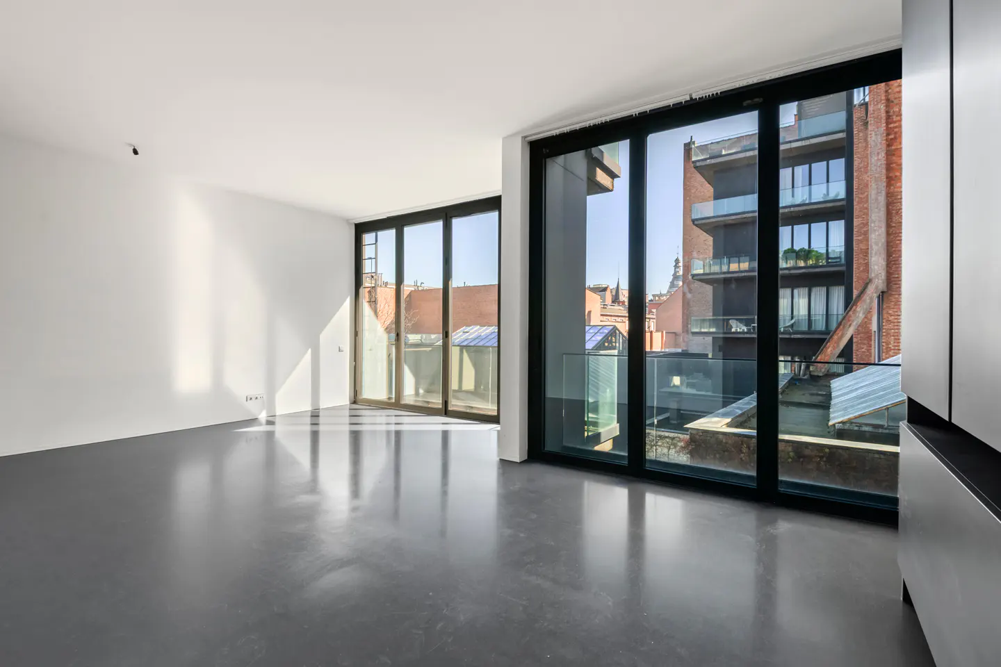 Bright, empty room with white walls, gray floor, and black-framed windows. Outside, buildings are visible.