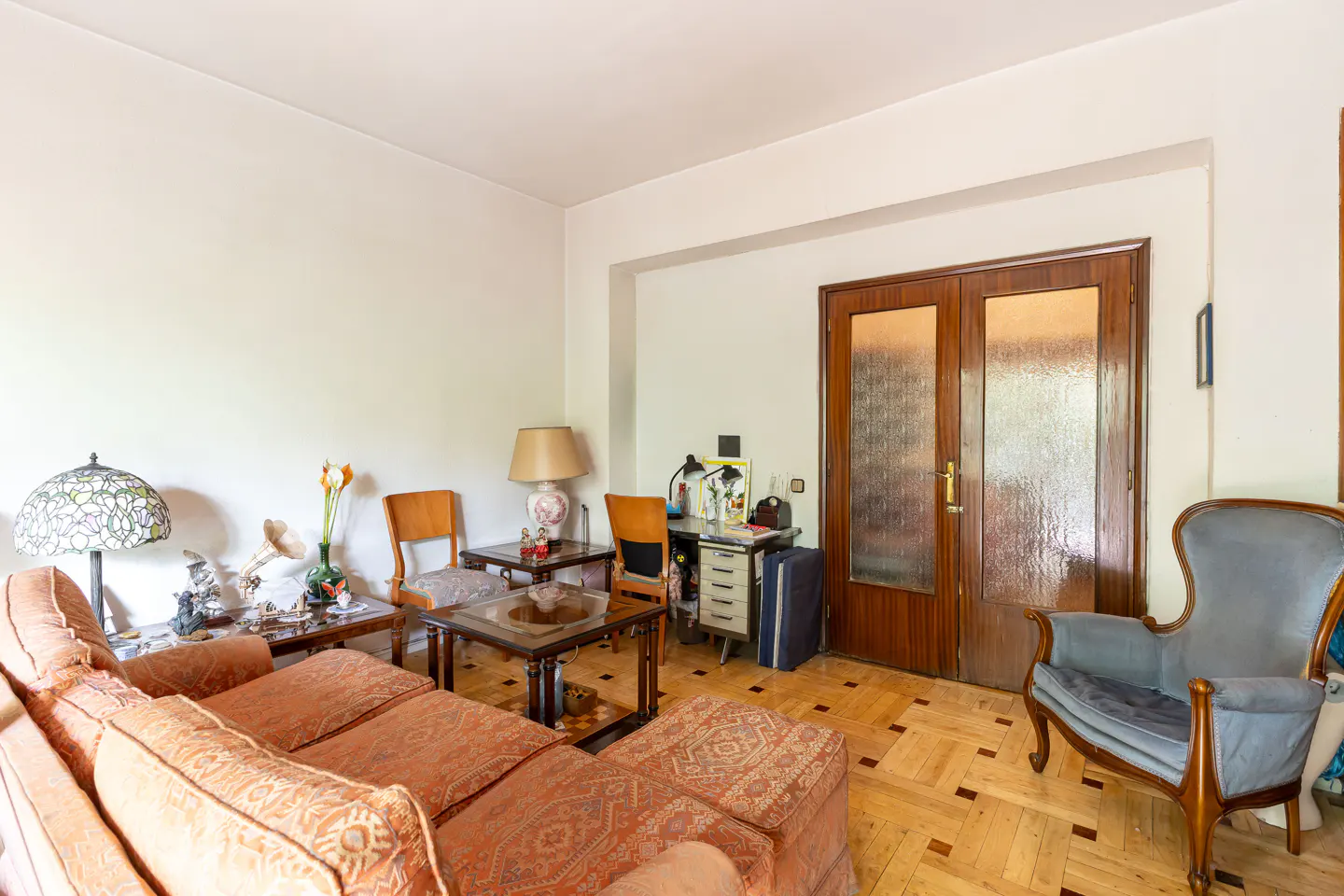 A living room with a patterned orange sofa, wooden chairs, and a blue armchair on a parquet floor.