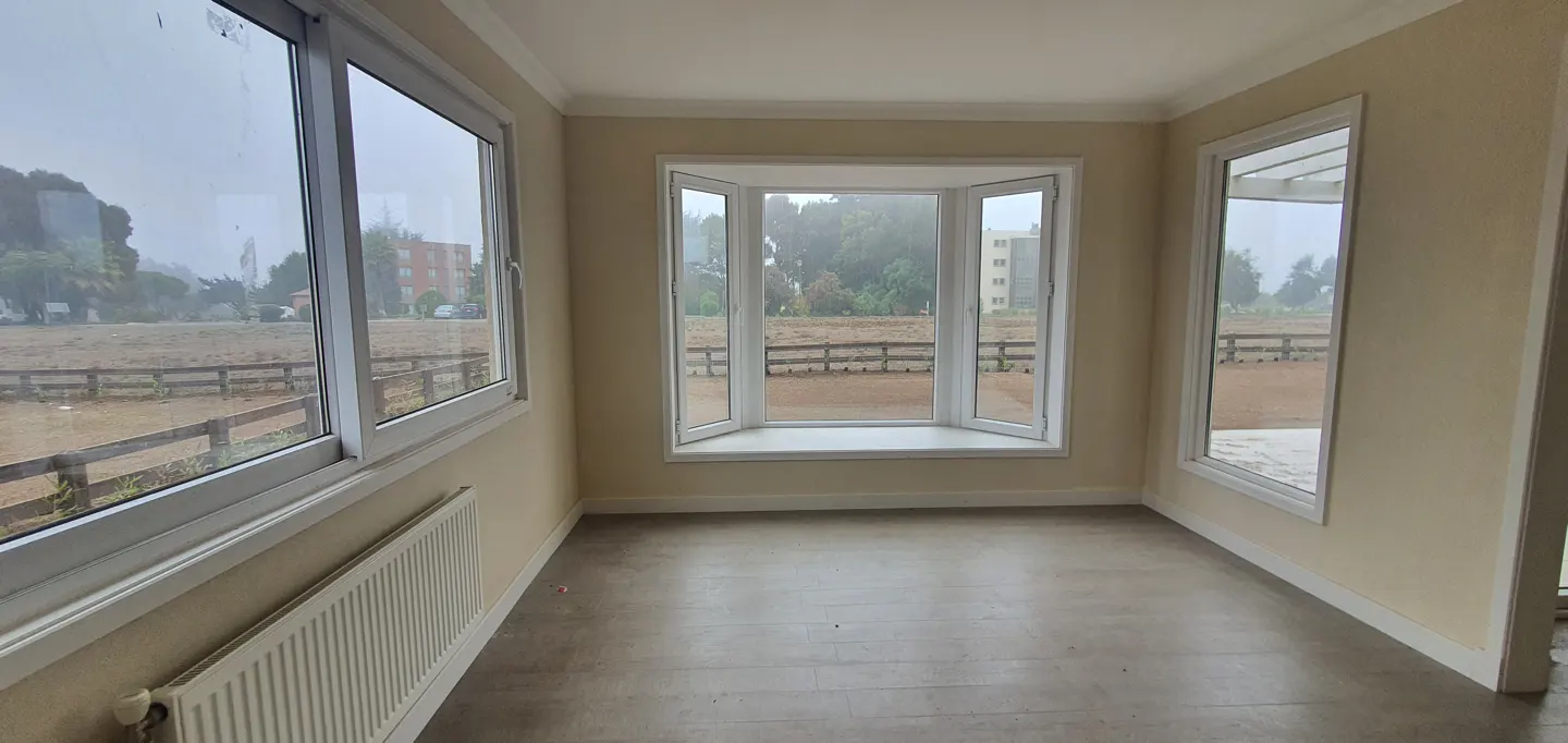 Empty room with beige walls, gray floor, and white trim. Large windows overlook a field with a wooden fence and trees.