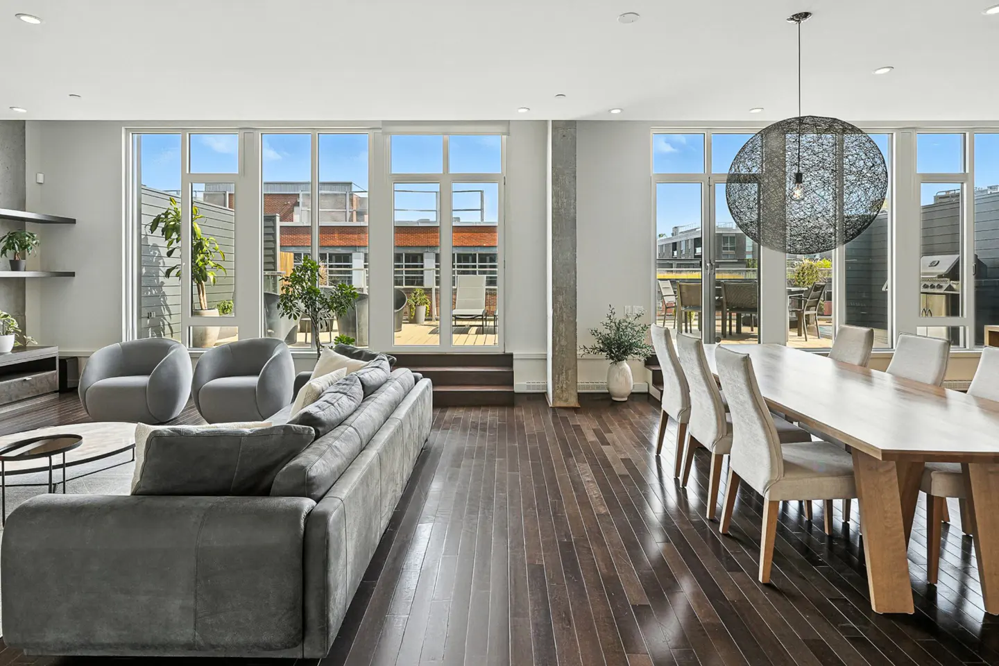 Bright, open-concept living and dining area with hardwood floors. Gray sofa, chairs, and a long wooden table with upholstered chairs. Large windows lead to a patio.