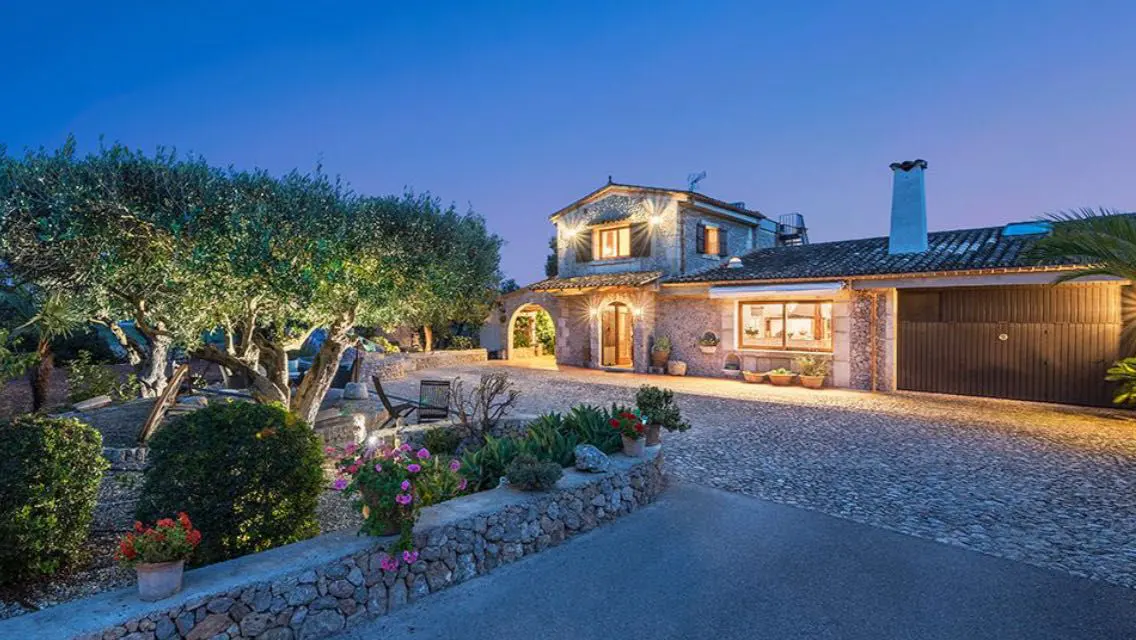 A stone house with lit windows and a stone driveway at dusk.