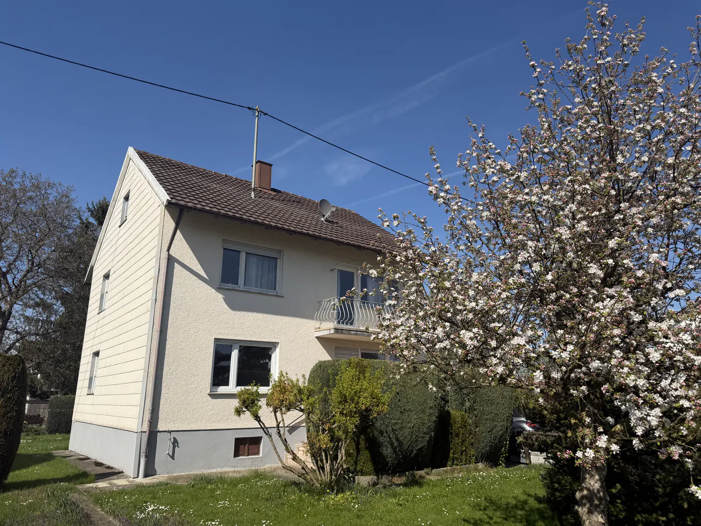 Two-story house with a brown roof, beige siding, and a small balcony, with a flowering tree in the yard.