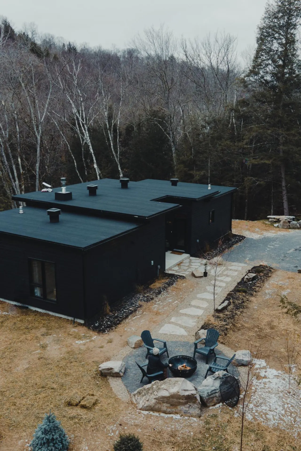 Modern black home with a flat roof, surrounded by trees. A stone path leads to a fire pit with chairs.