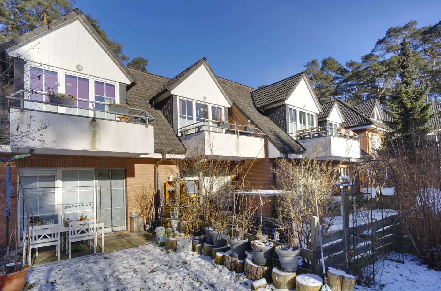 Row of brick townhouses with white gables and balconies under a blue sky. Snow covers the ground and potted plants line the patio.