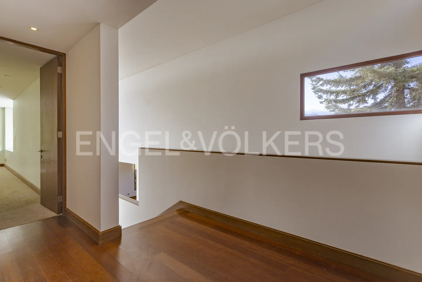 Hallway with wood floors, white walls, and a window showing a tree. The Engel & Völkers logo is visible on the wall.