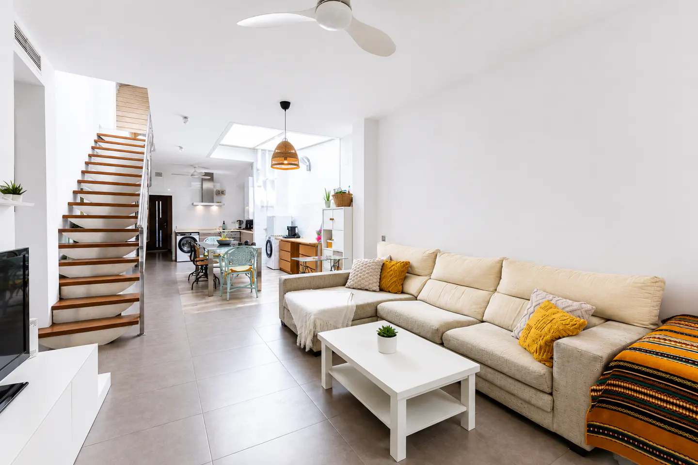 Modern open-plan living room with a beige sectional sofa, white coffee table, and wooden staircase leading to a kitchen.