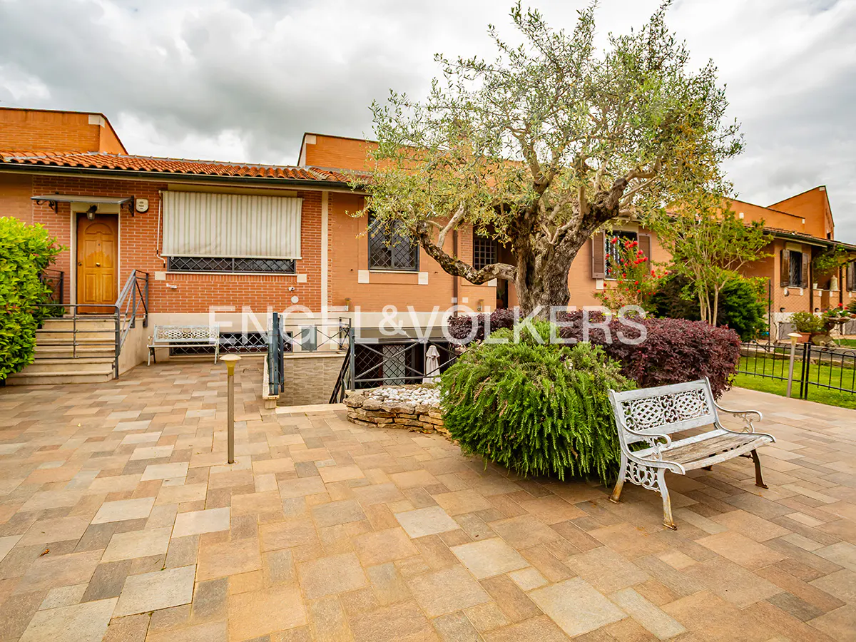 Exterior view of a brick house with a stone patio, a white bench, and a large tree. The Engel & Völkers logo is visible.