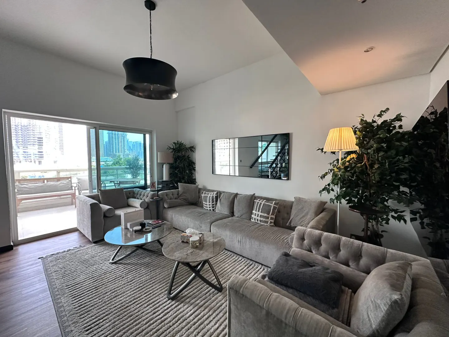 Living room with gray sectional sofa, glass coffee tables, and a balcony with city views. A black pendant light hangs above.