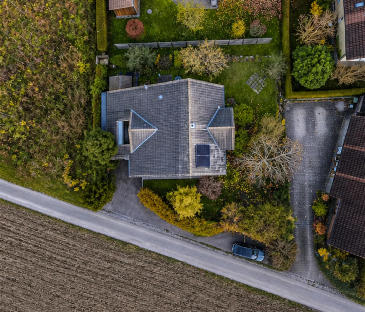 Aerial view of a house with a gray roof, solar panels, and a well-maintained garden in autumn. A road and a field are visible.