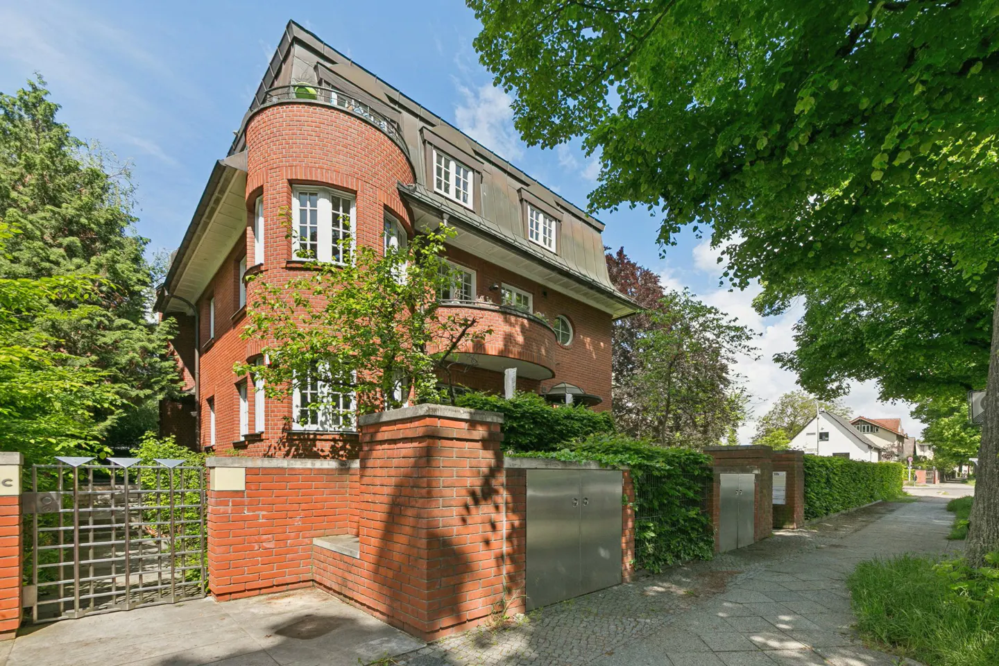 Exterior view of a three-story red brick house with white-framed windows, a curved corner, and a dark roof, surrounded by green trees and a brick fence.