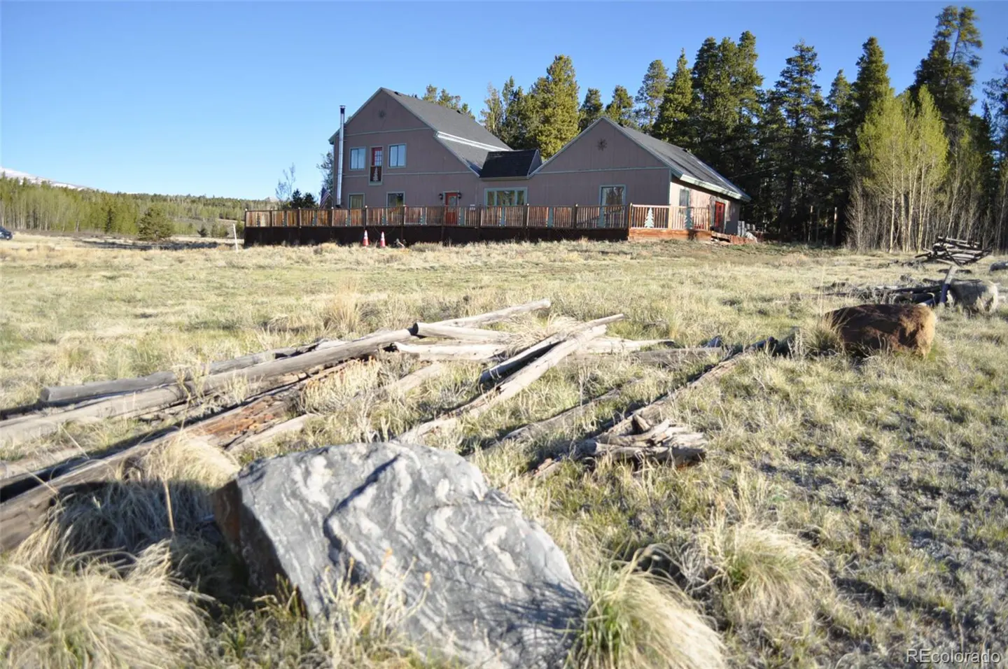A two-story tan house with a large wooden deck sits in a grassy field with scattered logs and rocks, backed by pine trees.