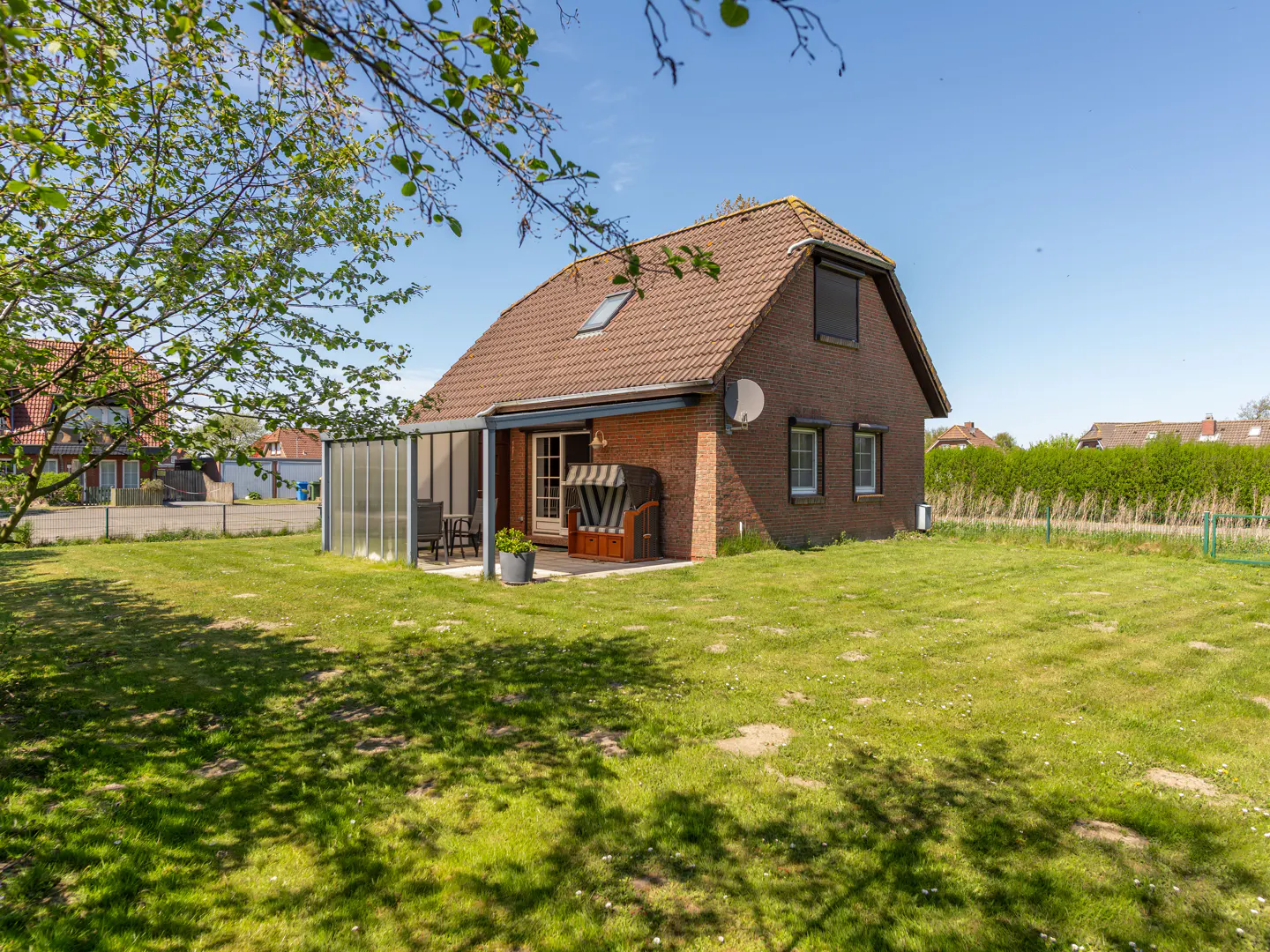 A brick house with a brown tiled roof, a patio with a beach chair, and a large green lawn under a clear blue sky.