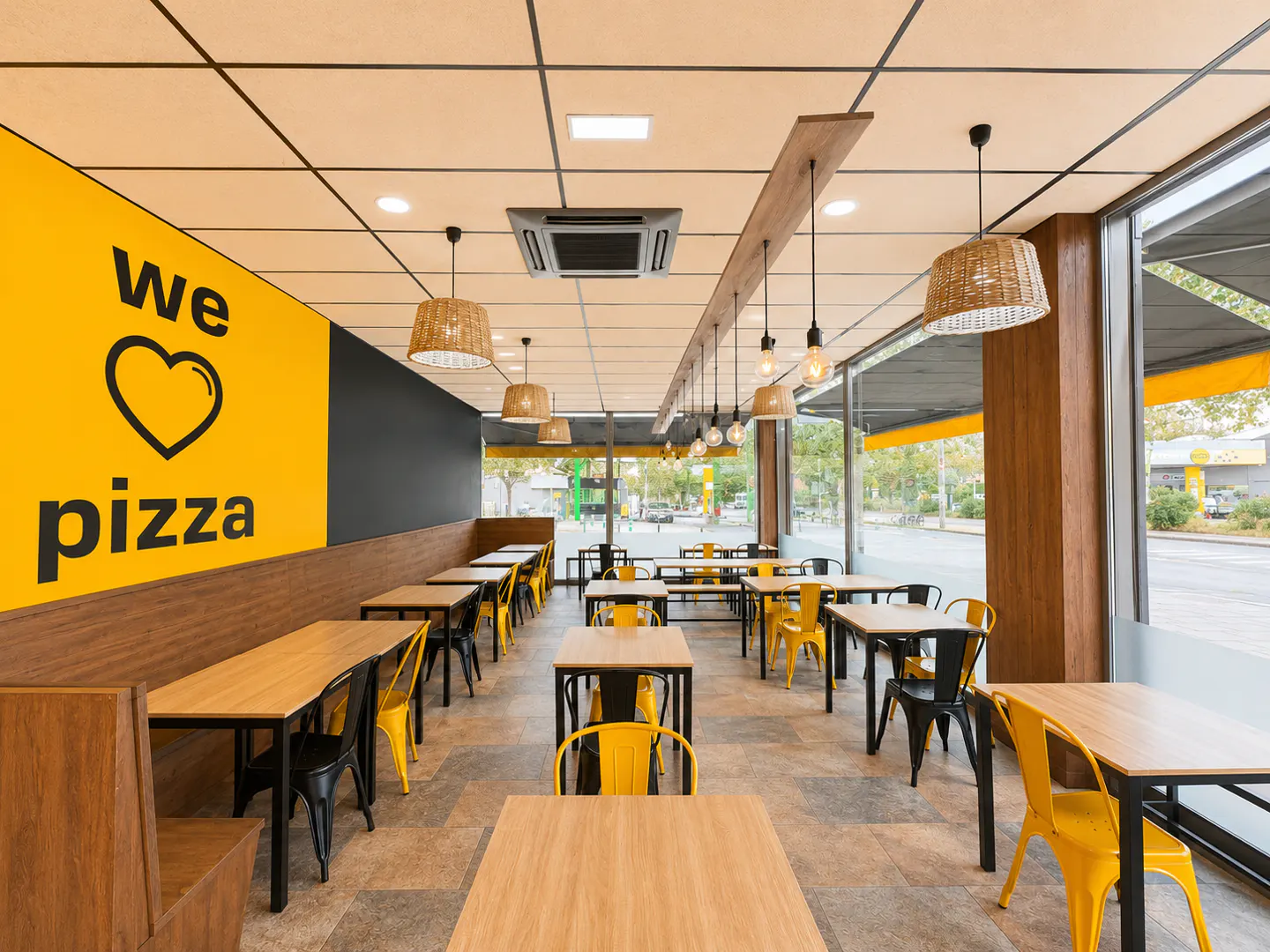 Modern pizza restaurant interior with yellow walls, wooden tables, black and yellow chairs, and hanging wicker lights.