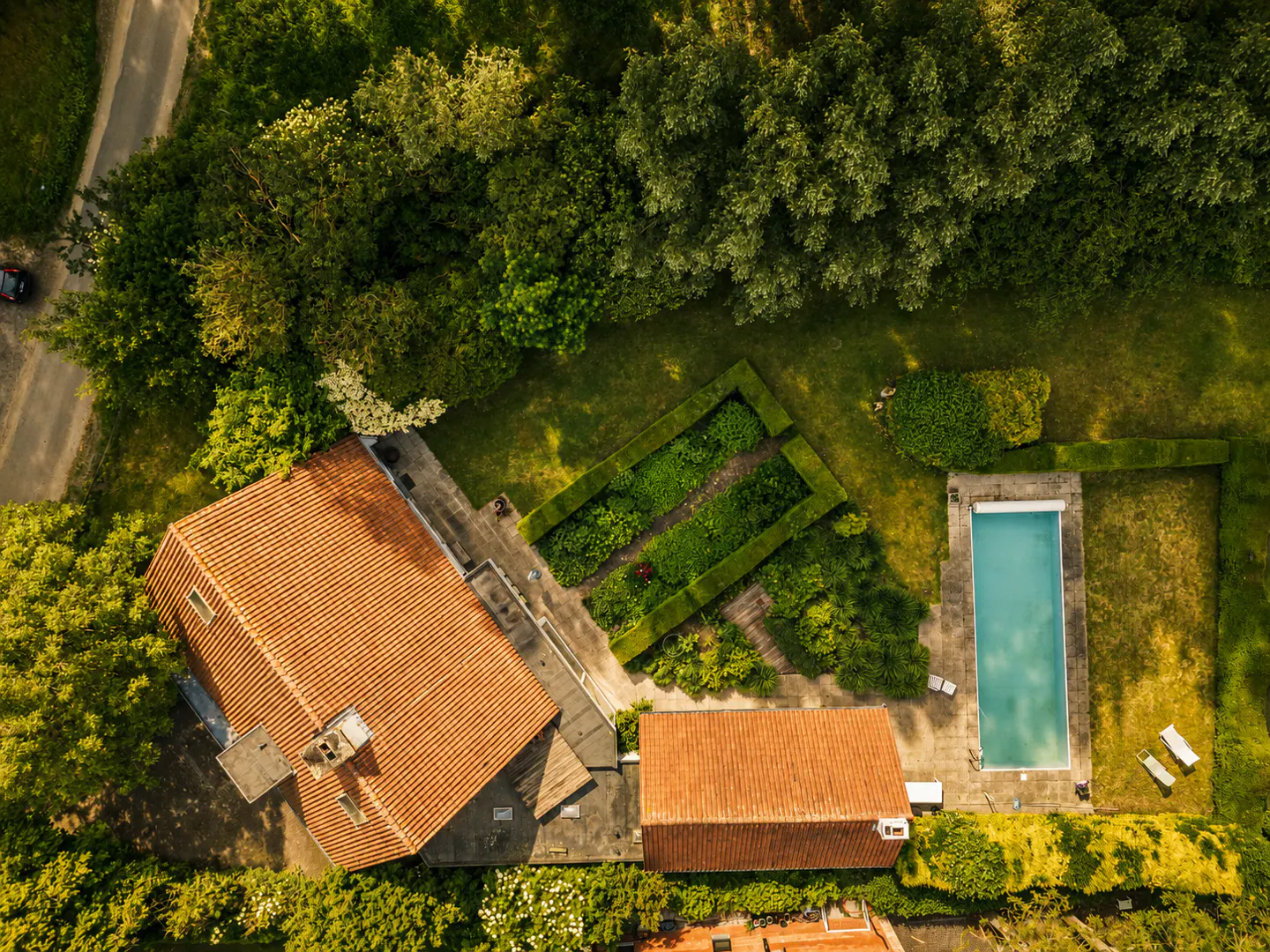 Aerial view of a house with a terracotta roof, a rectangular swimming pool, and a manicured garden surrounded by trees.