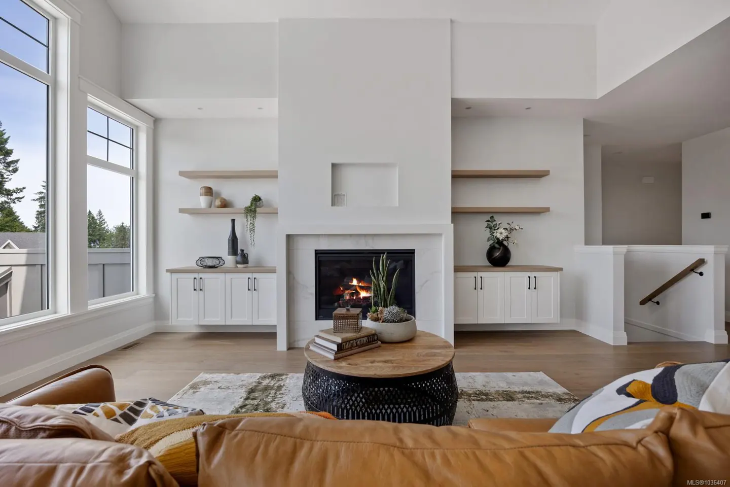 Bright living room with a tan leather couch, a black coffee table, and a white fireplace with a burning fire. Built-in shelves and cabinets flank the fireplace. Large windows let in natural light.