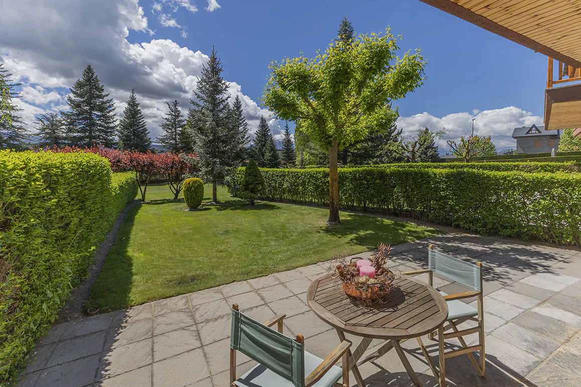 Patio with a round wooden table, two chairs, and a basket centerpiece. Green lawn, trees, and blue sky with clouds in the background.