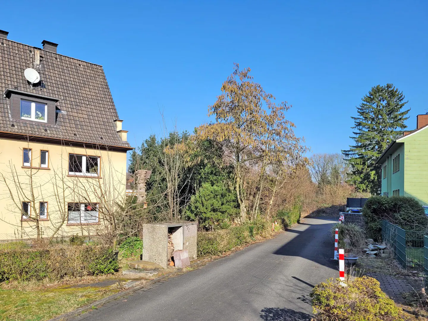 Street view of a yellow house with a satellite dish, trees, and a concrete structure on a sunny day.