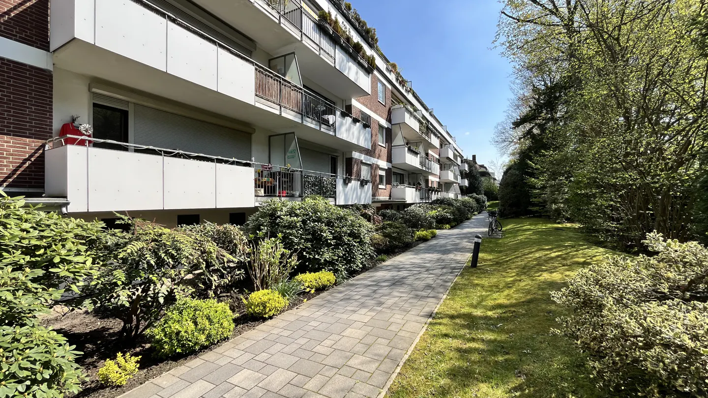 Brick apartment building with white balconies, lush green bushes, and a paved walkway on a sunny day.