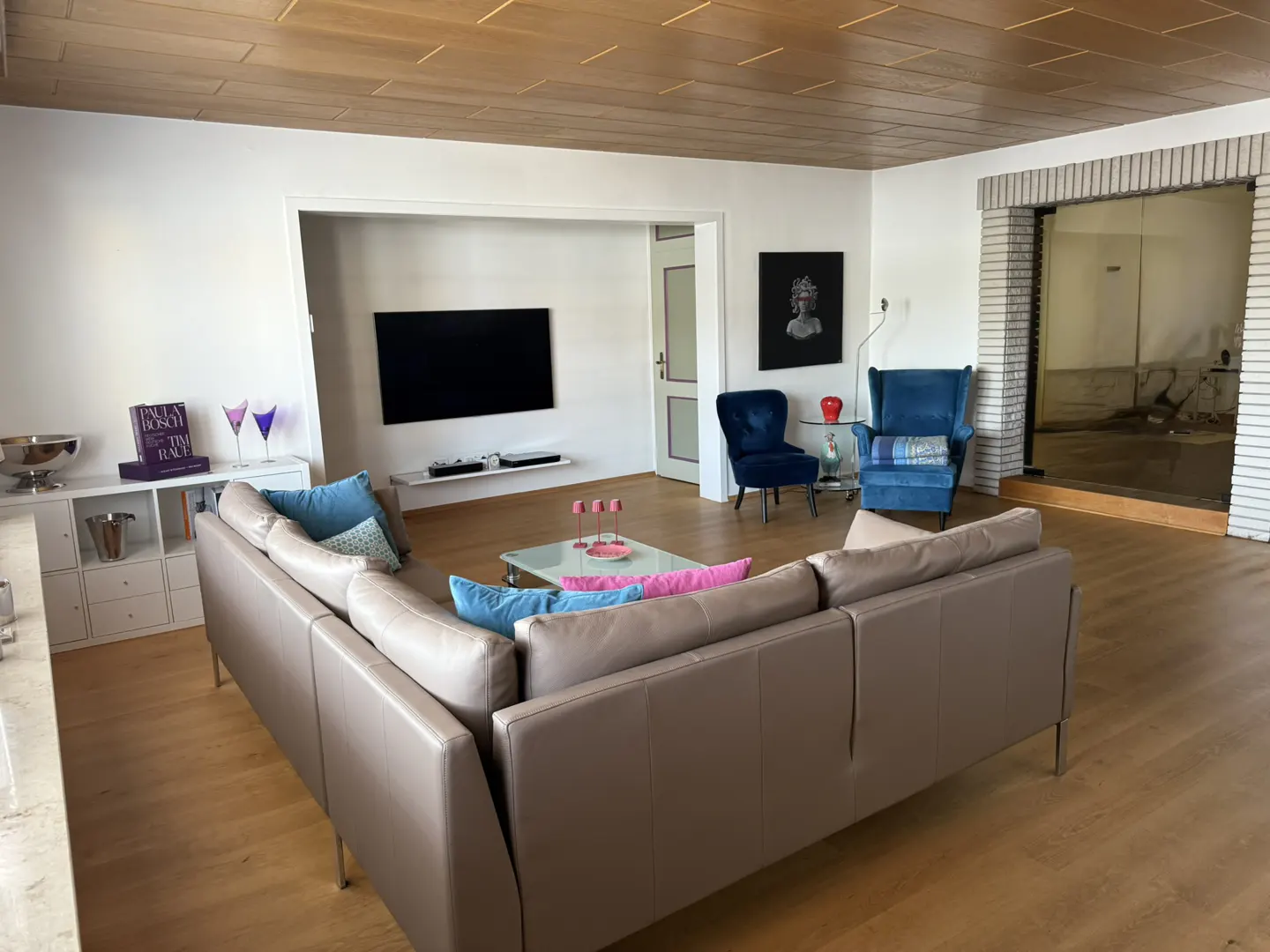 Living room with a brown leather sofa, blue chairs, and a TV. The room has a wood floor and a wood ceiling.