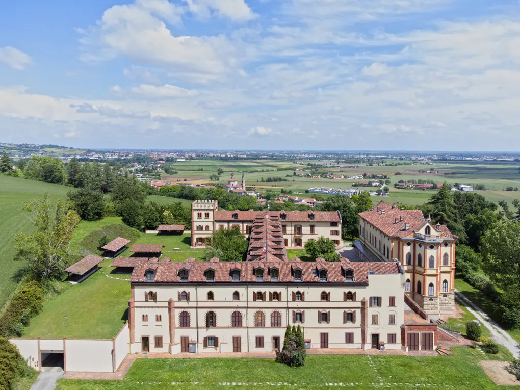 Aerial view of a large, light-yellow building with a red-tiled roof, surrounded by green grass and trees, with a town in the distance.