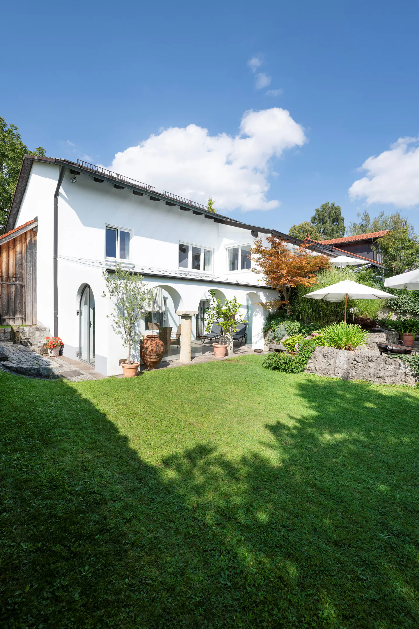 Exterior of a white house with arched doorways, a green lawn, and patio furniture under white umbrellas.