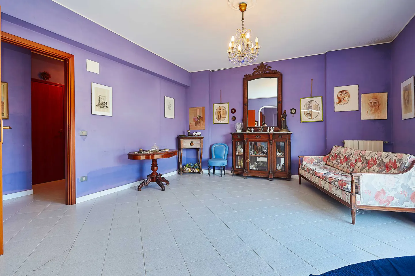 A living room with purple walls, a floral sofa, a wooden cabinet with a mirror, and a chandelier.