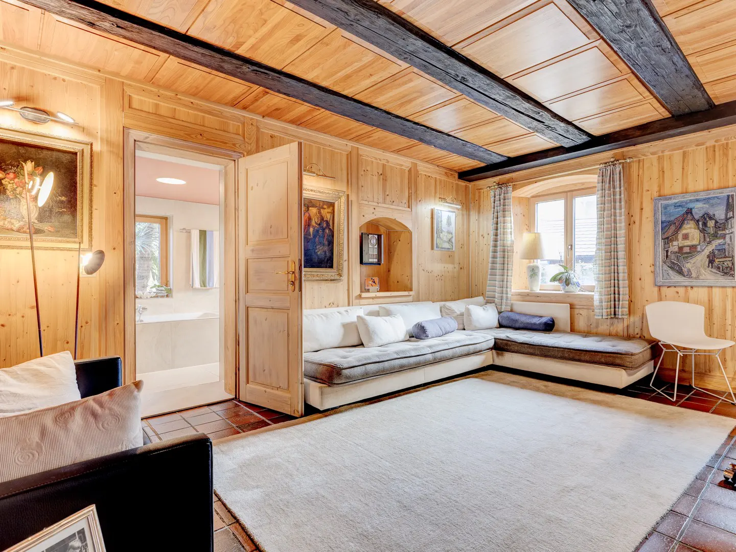 Living room with wood paneled walls, dark wood beams, and a white L-shaped sofa. A bathroom is visible through an open door.