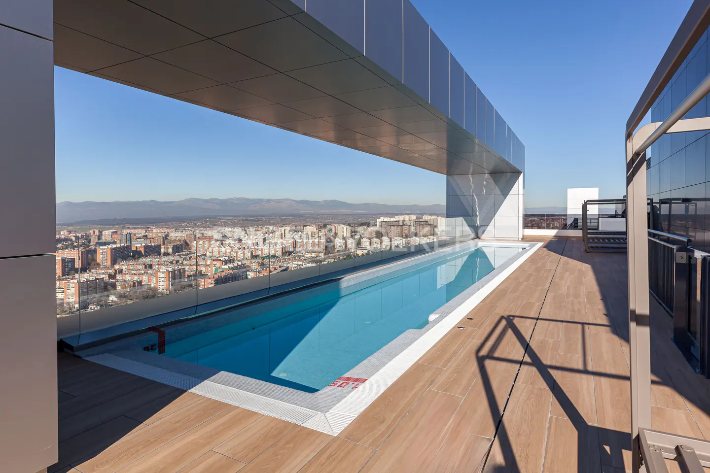 Rooftop pool with blue water and wood deck overlooks a city skyline under a clear blue sky. Modern architecture frames the view.