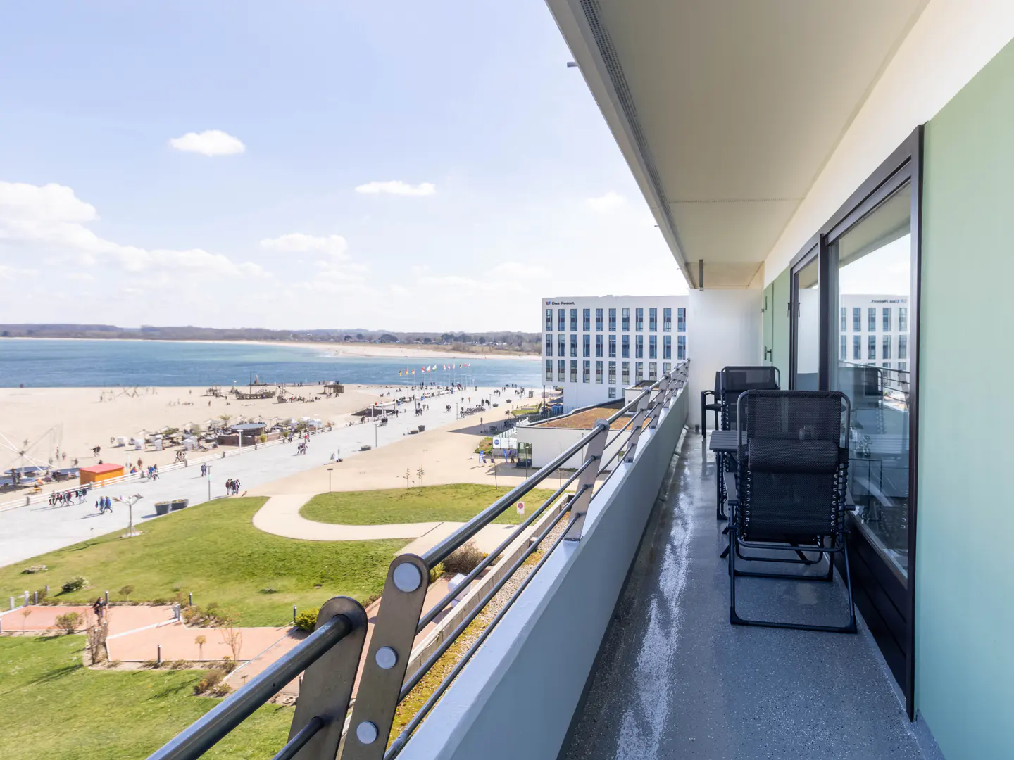 Balcony view of a beach. Black chairs sit on a grey balcony overlooking a sandy beach, blue water, and a white building under a blue sky.