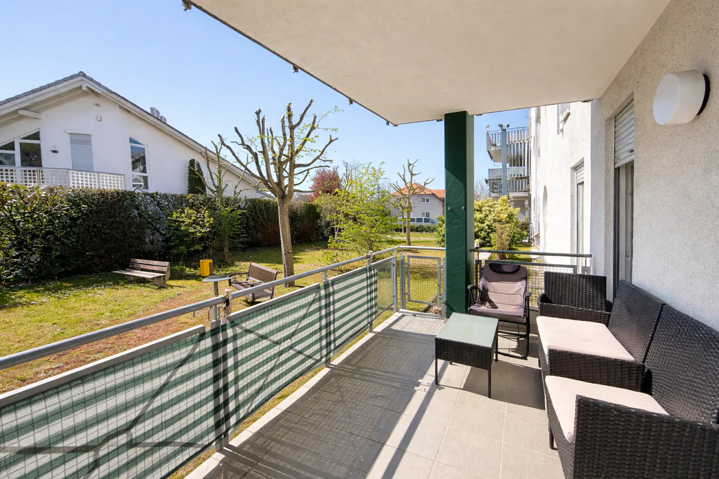 Balcony with wicker furniture overlooks a green lawn with benches and trees on a sunny day.
