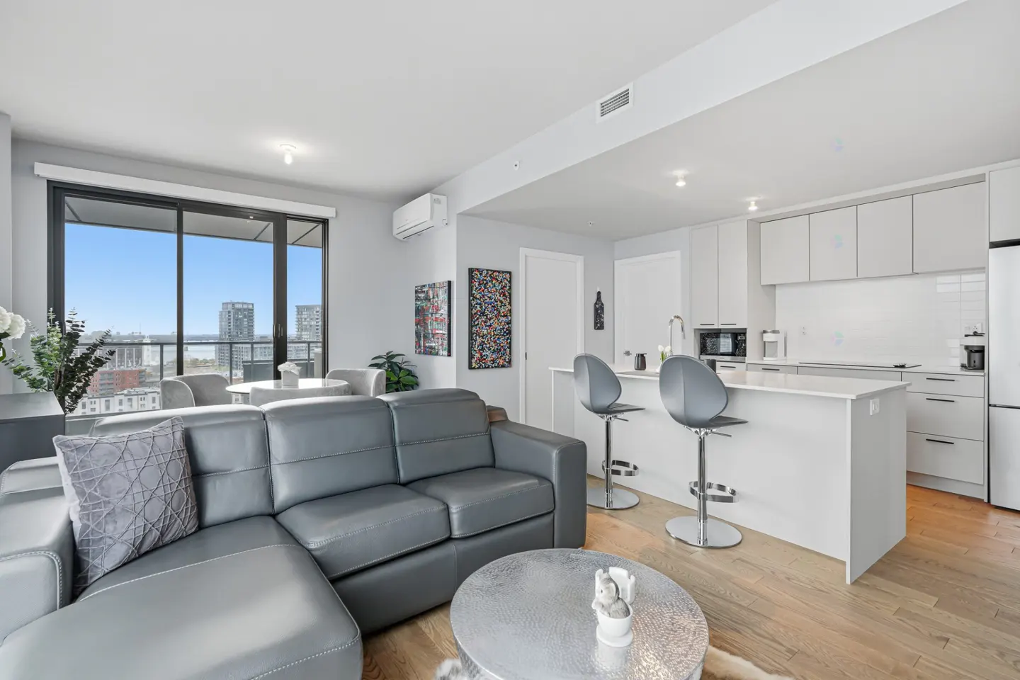 Bright, modern condo interior with gray leather sofa, white kitchen island, and city view from large windows.