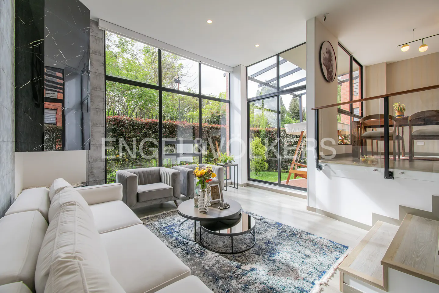 Bright living room with white sofa, grey chairs, and large windows overlooking a green garden. A black marble fireplace adds a modern touch.