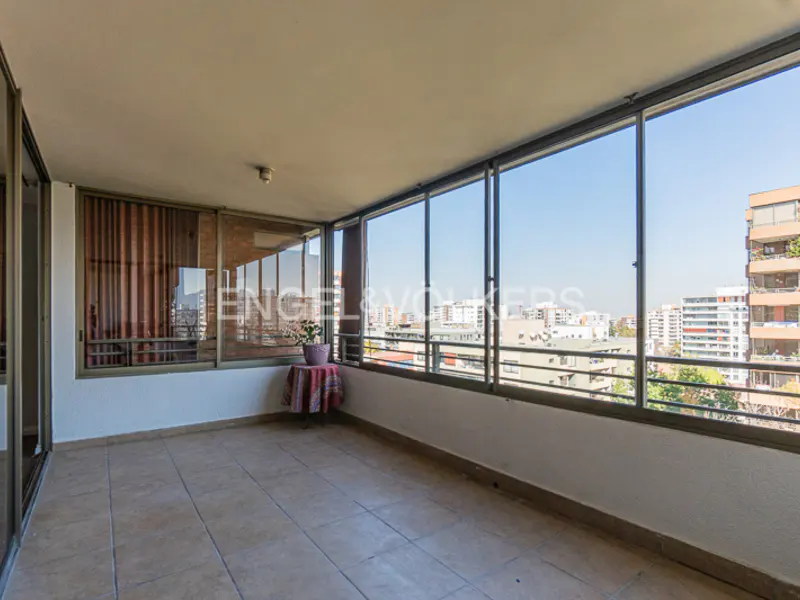A sunroom with large windows overlooking a city skyline. A small table with flowers sits near the windows. The floor is tiled.