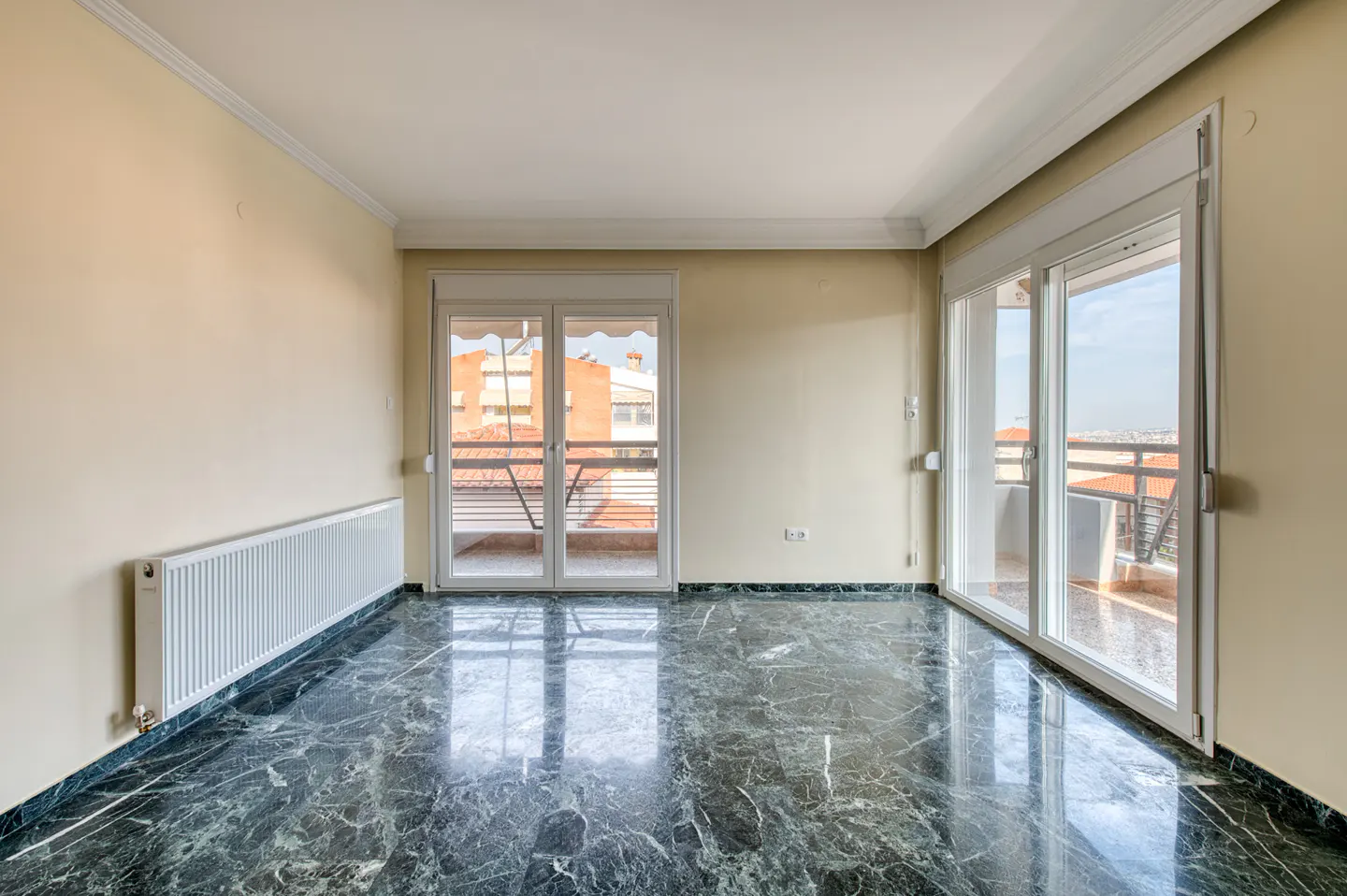 Empty room with marble floor, cream walls, and white trim. Two sets of sliding glass doors lead to balconies. A white radiator is on the left wall.