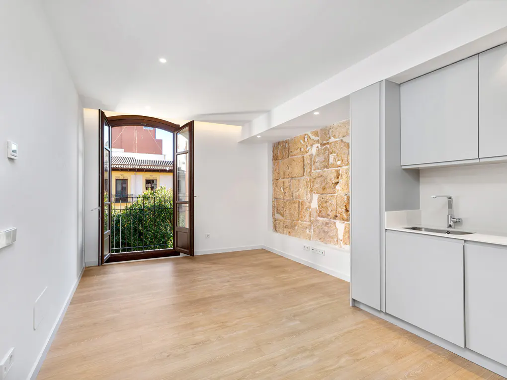 Bright, empty room with wood floors, white walls, and a stone accent wall. Open balcony doors show greenery and a building. Gray kitchen cabinets are on the right.