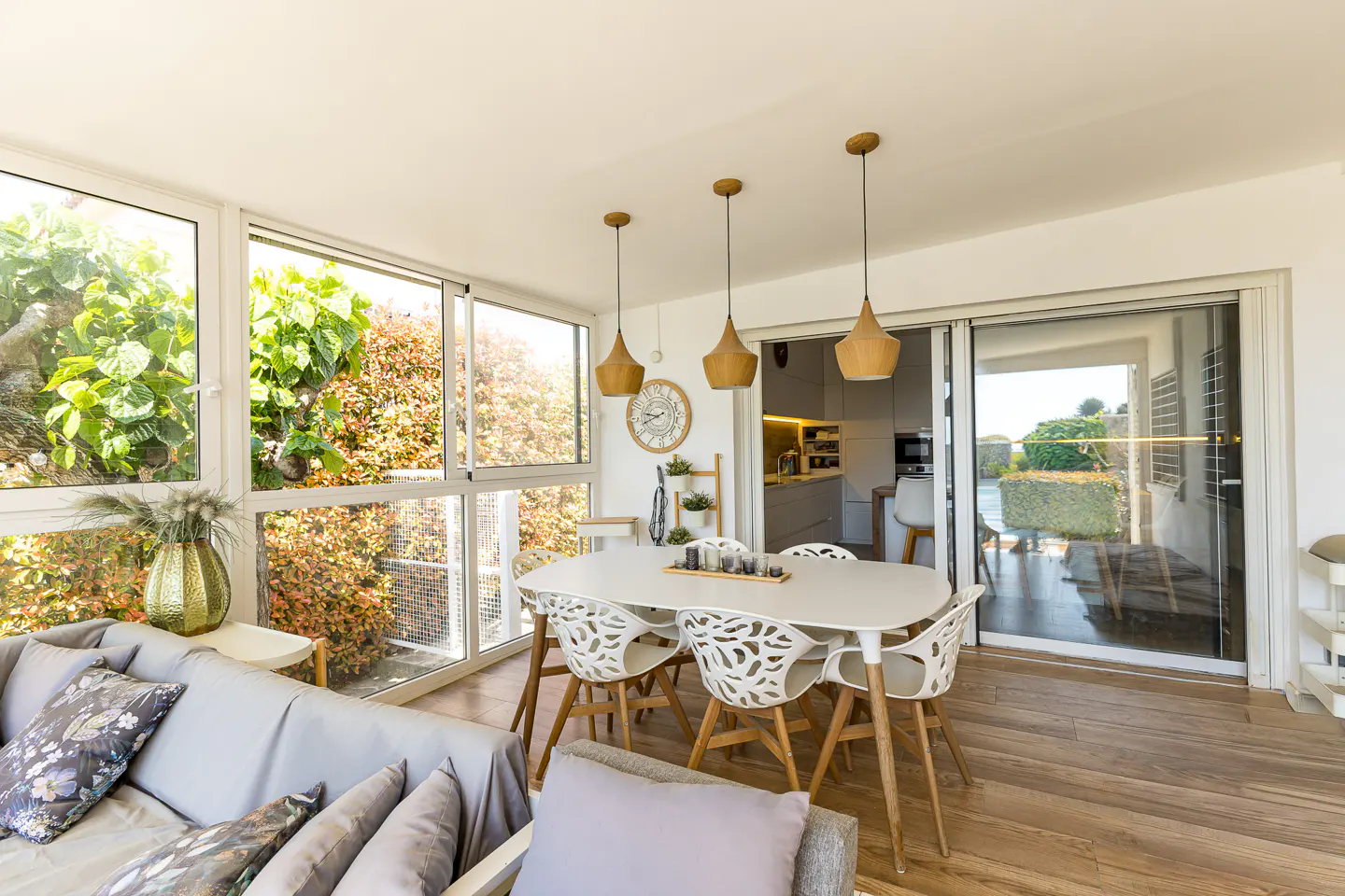 Bright sunroom with a gray sofa, white table and chairs, and wood floors. Large windows overlook lush greenery. Three pendant lights hang above the table.