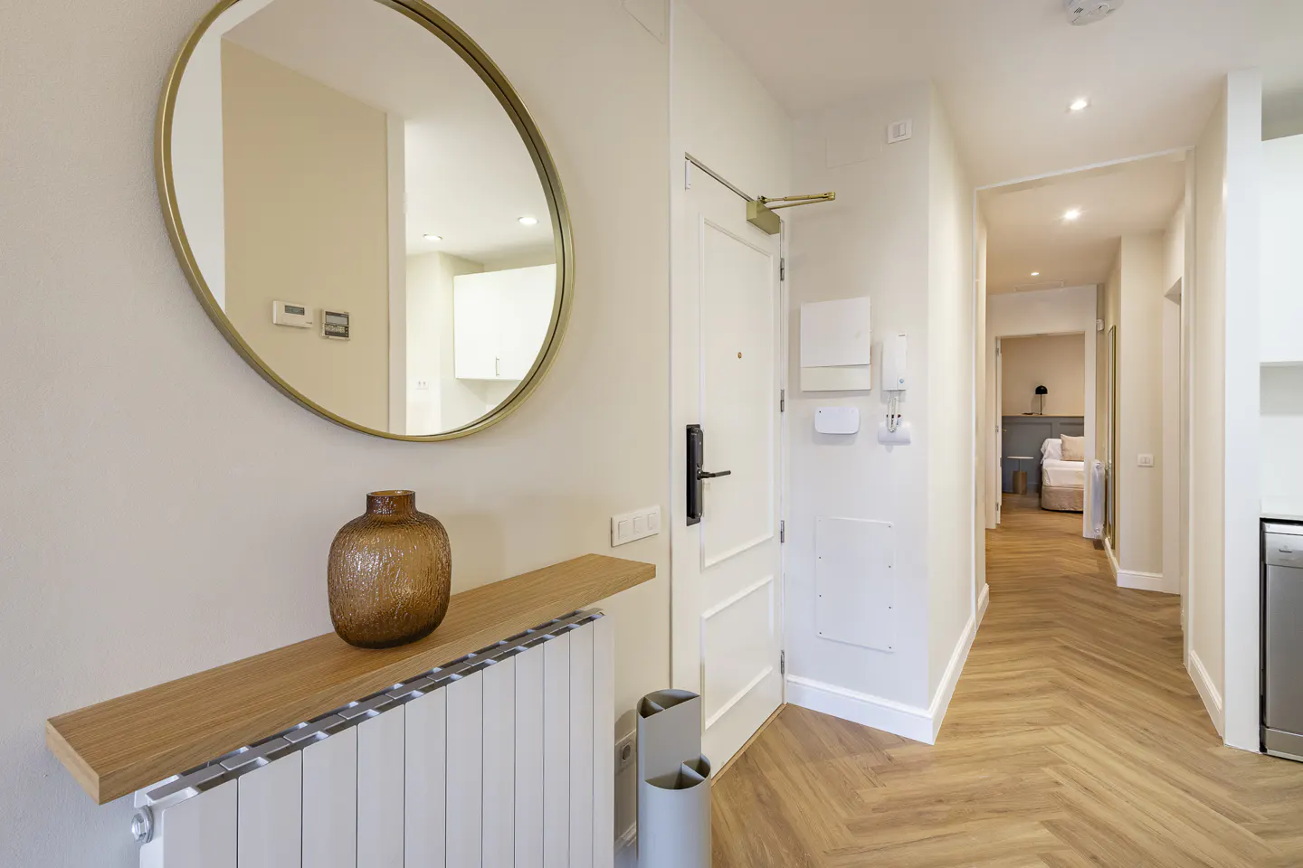Apartment entryway with a round mirror, brown vase on a wooden shelf above a radiator, and a hallway leading to a bedroom.