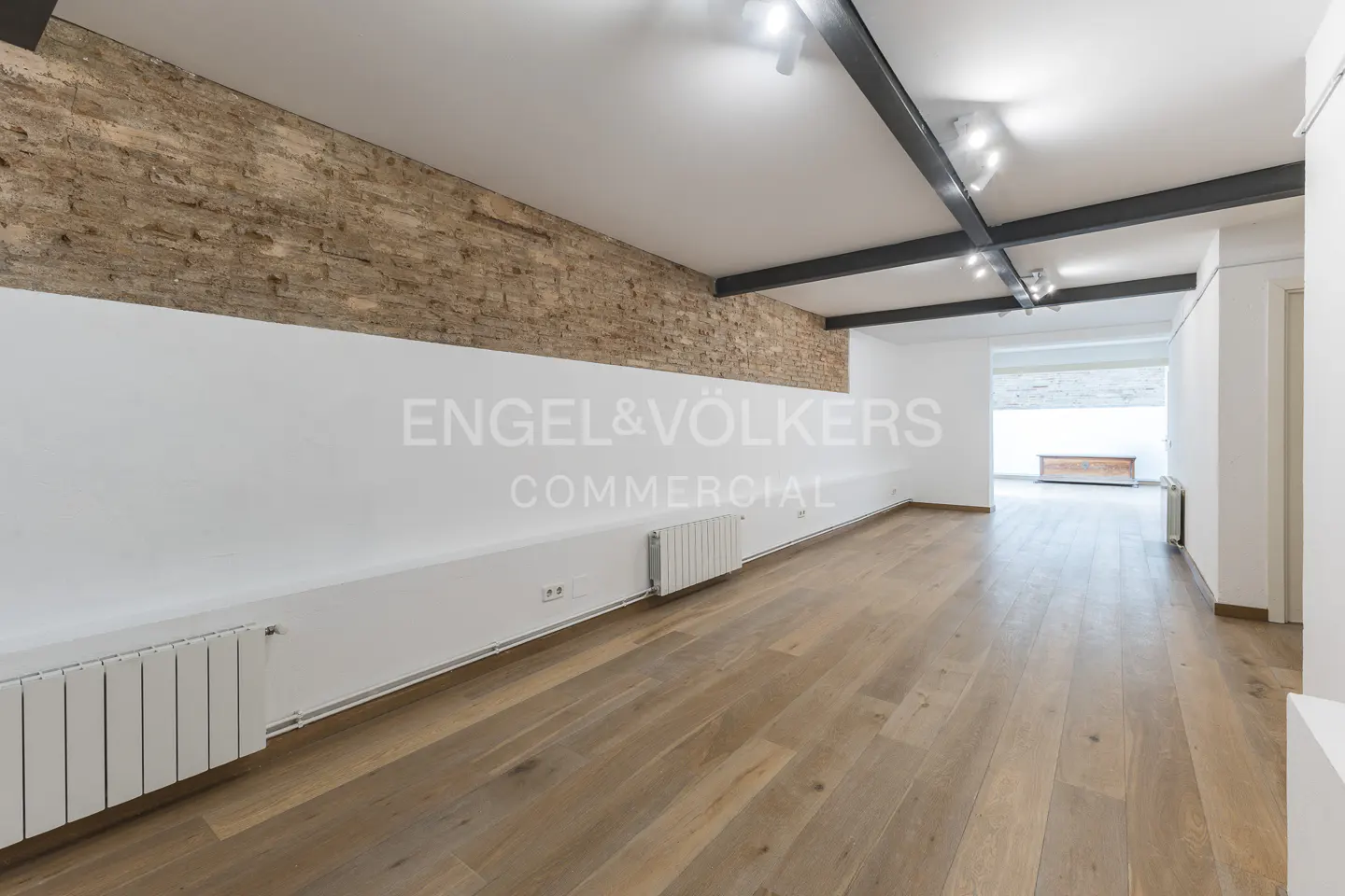 Empty commercial space with hardwood floors, white walls, and a brick accent wall. Black beams and track lighting on the ceiling.