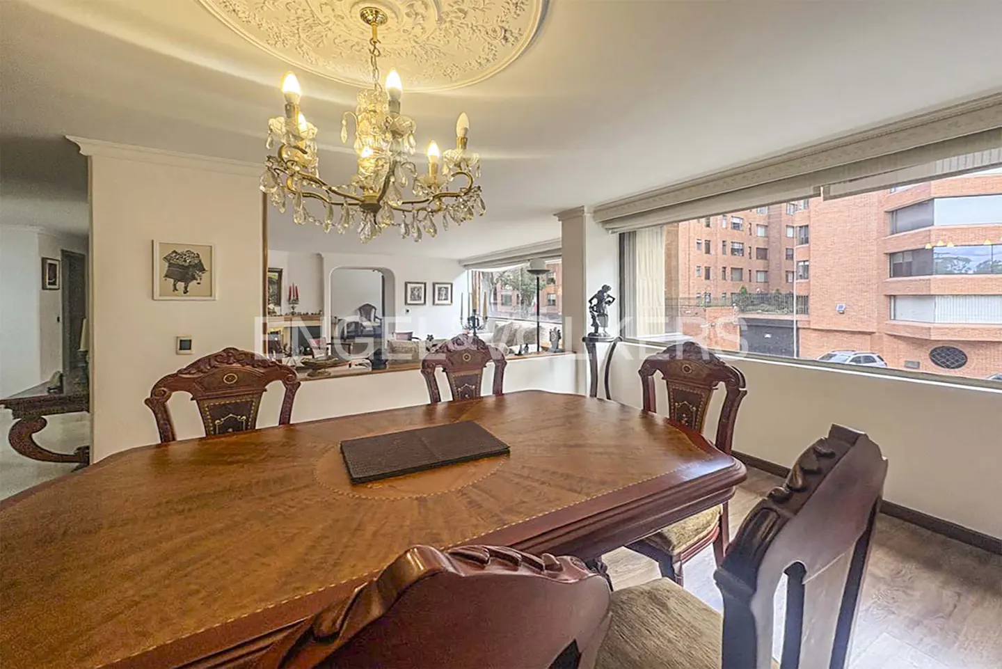 A dining room with a large wooden table and chairs under a chandelier. A window shows a brick building outside.