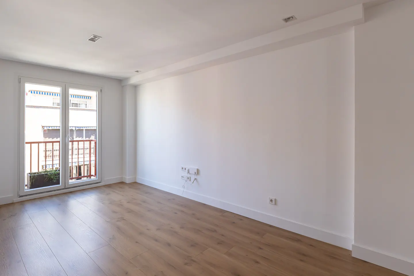 Bright, empty room with light wood floors, white walls, and a balcony visible through a glass door.