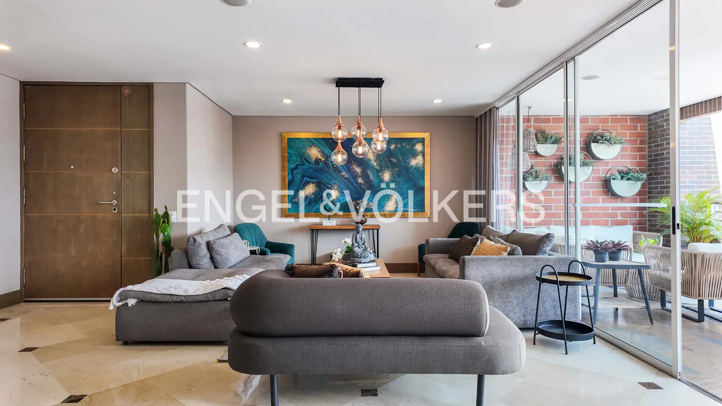 A modern living room with gray sofas, a blue and gold painting, and a glass-enclosed balcony with brick wall and plants.
